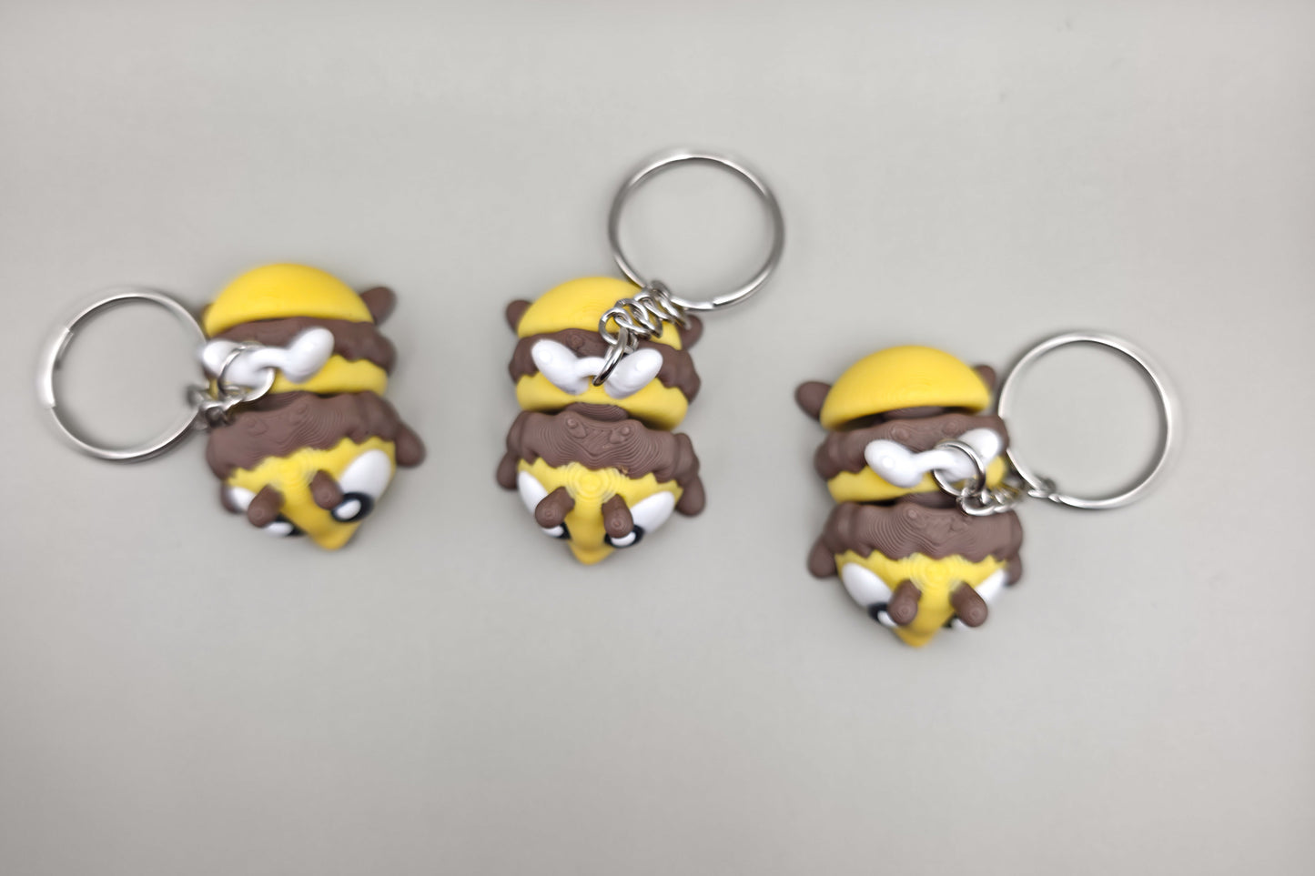 Trois porte-clés Abeille articulés MinoZoo jaune, brun et blanc, présentés de dessus sur fond gris.