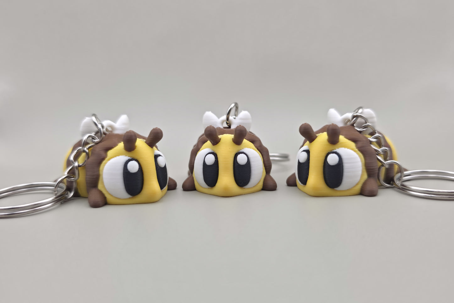 Trois porte-clés Abeille articulés MinoZoo jaune, brun et blanc, alignés de face sur fond gris.