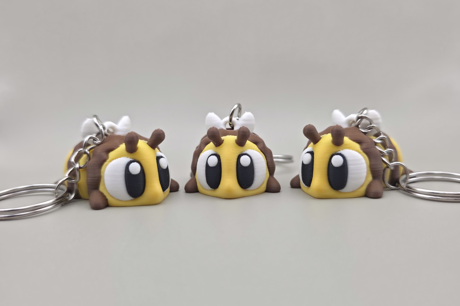 Trois porte-clés Abeille articulés MinoZoo jaune, brun et blanc, alignés de face sur fond gris.