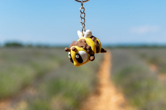 Porte-clés Abeille articulé MinoZoo jaune, brun et blanc, suspendu de trois quarts par sa chaîne au-dessus d’une prairie.
