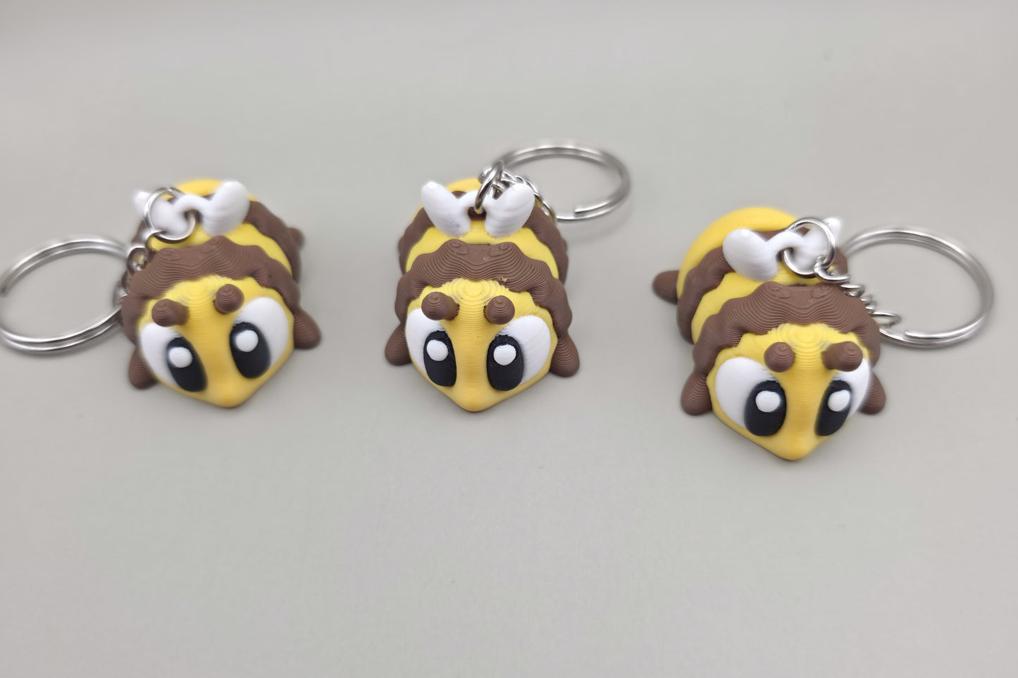 Trois porte-clés Abeille articulés MinoZoo jaune, brun et blanc, disposés de trois quarts sur fond gris.