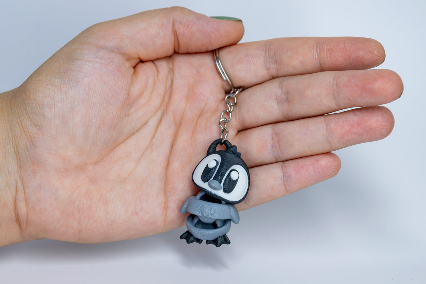Porte-clés Bébé Pingouin articulé MinoZoo noir, blanc et gris moyen, tenu de face dans une main avec sa chaîne.