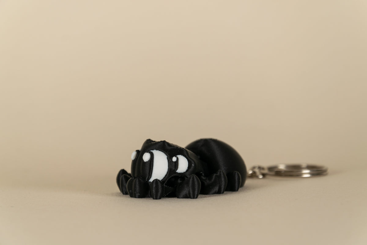 Porte-clés Black Spider articulé MinoZoo noir et blanc, vue trois-quarts sur fond beige.