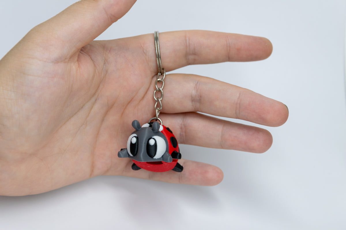 Porte-clés Coccinelle articulé MinoZoo rouge, gris foncé et noir, tenu dans une main en vue trois quarts.