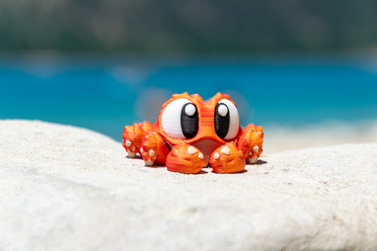 Porte-clés Crabe articulé MinoZoo aux reflets orange à rouge, posé de face sur un rocher au bord de l’eau.
