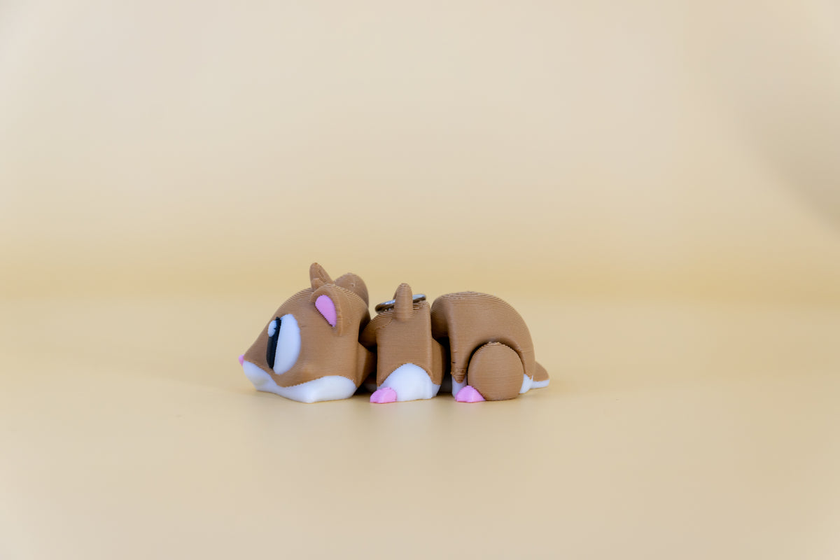 Porte-clés Hamster articulé MinoZoo brun, blanc et rose, vue de profil sur fond beige.