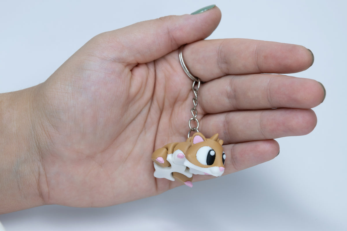 Porte-clés Hamster articulé MinoZoo brun, blanc et rose, vue trois-quarts tenu dans une main.