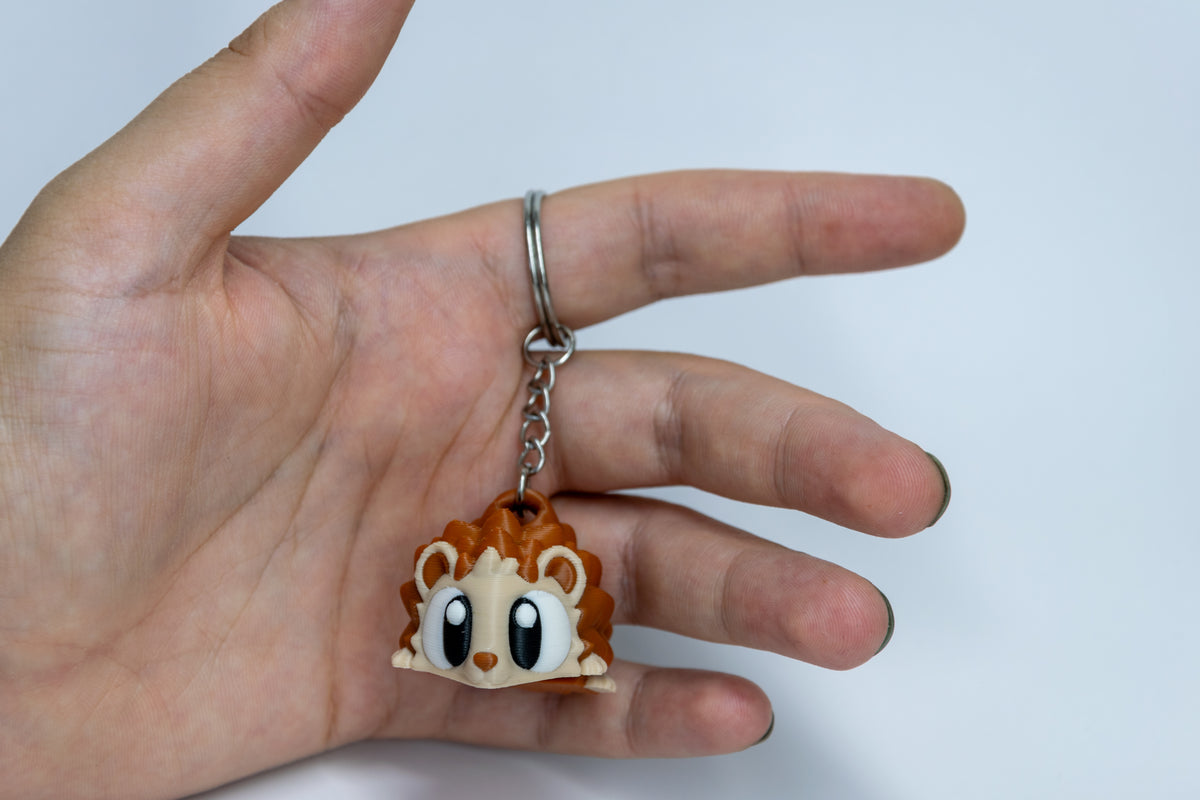 Porte-clés Hérisson articulé MinoZoo brun et beige, tenu de face dans une main avec chaîne et anneau visibles.