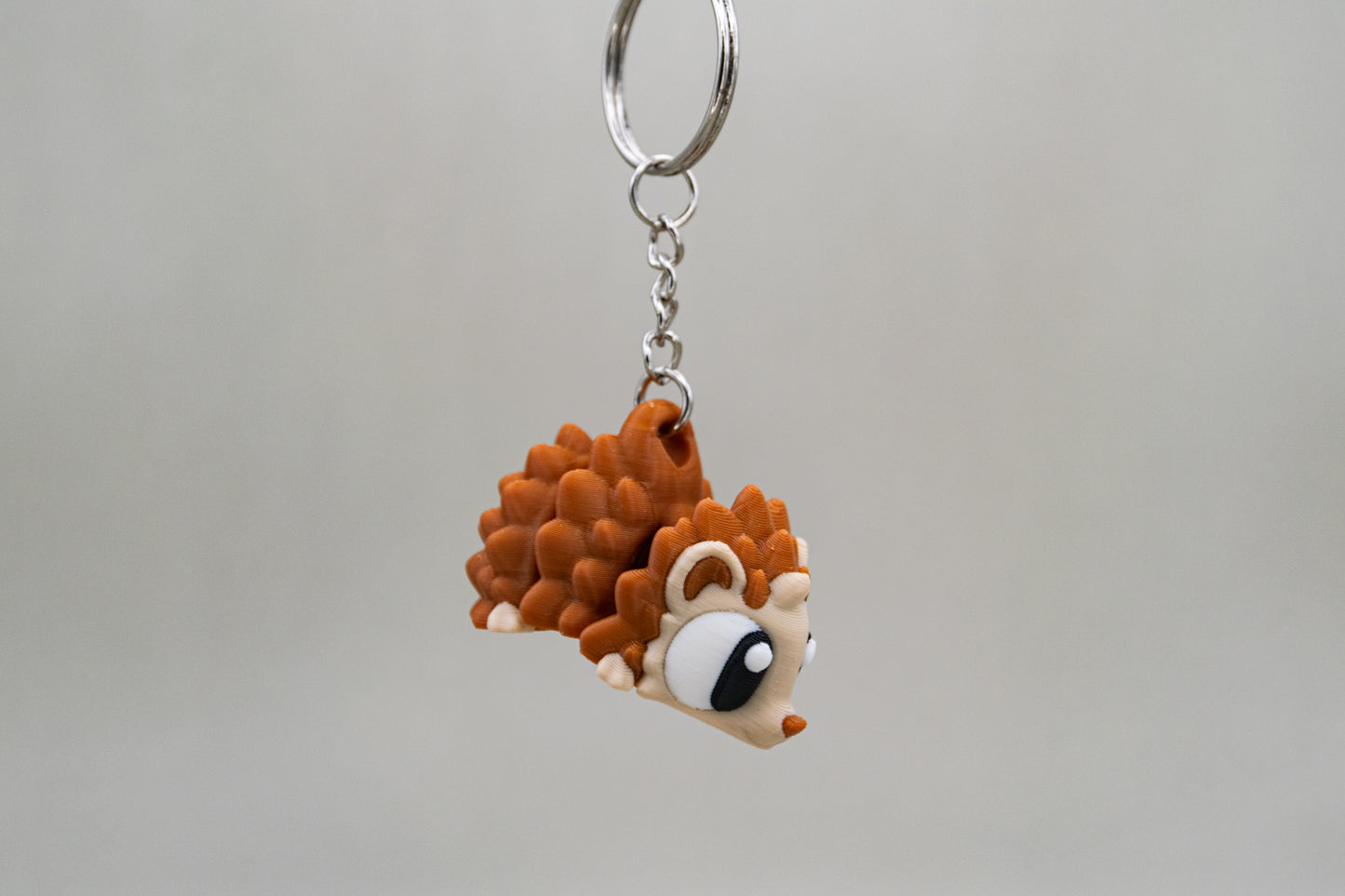 Porte-clés Hérisson articulé MinoZoo beige et brun caramel, suspendu de profil sur fond gris.