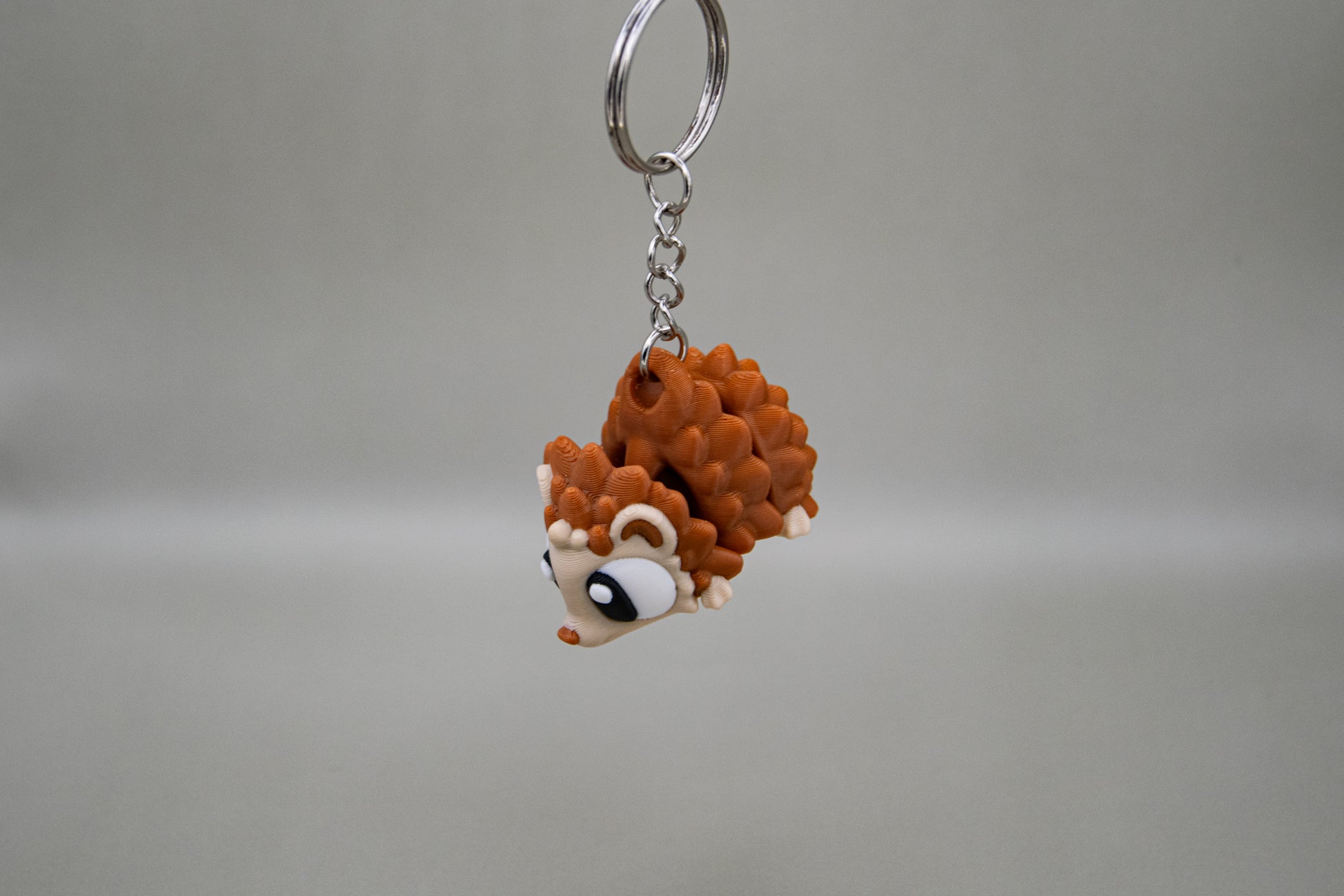 Porte-clés Hérisson articulé MinoZoo beige et brun caramel, suspendu de trois quarts sur fond gris.