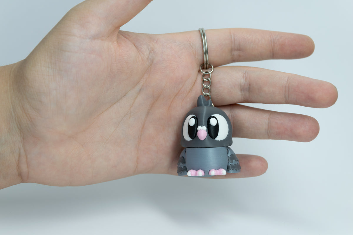 Porte-clés Pigeon articulé MinoZoo gris foncé, gris bleuté et rose, tenu de face dans une main.