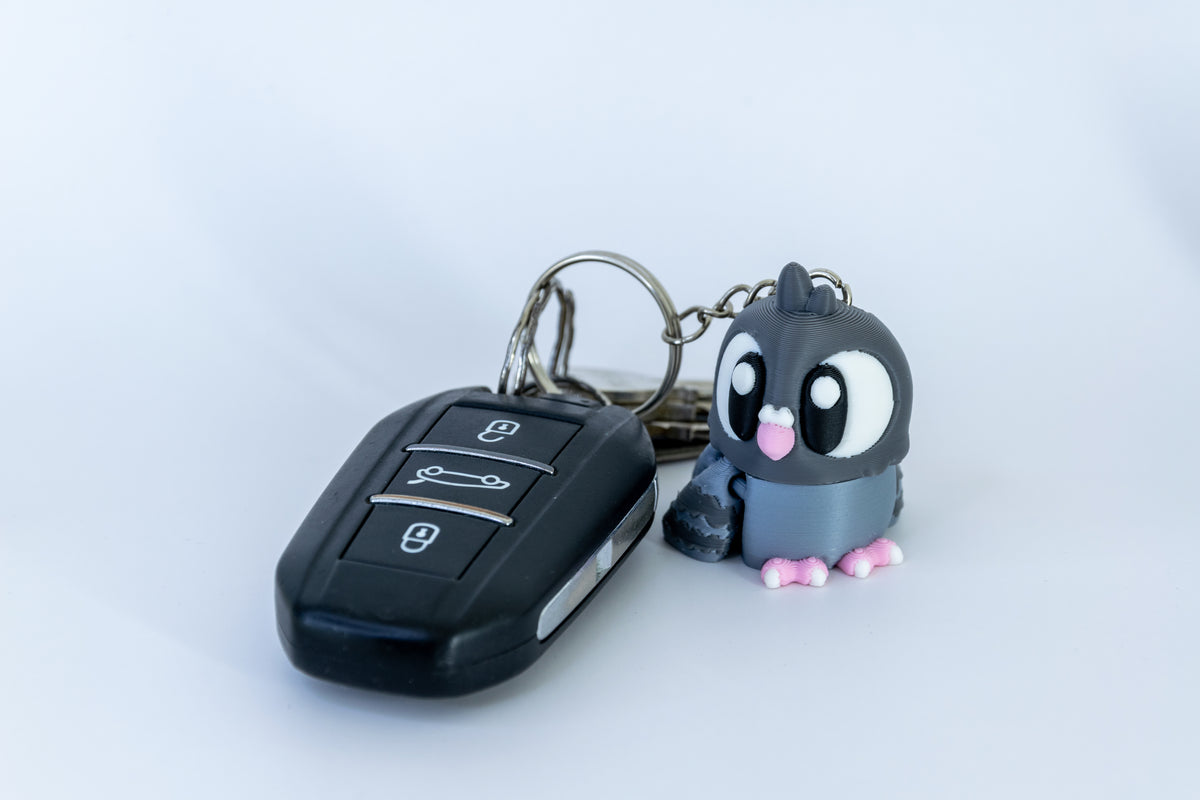 Porte-clés Pigeon articulé MinoZoo gris foncé et gris bleuté, de trois quarts près d’une clé de voiture.